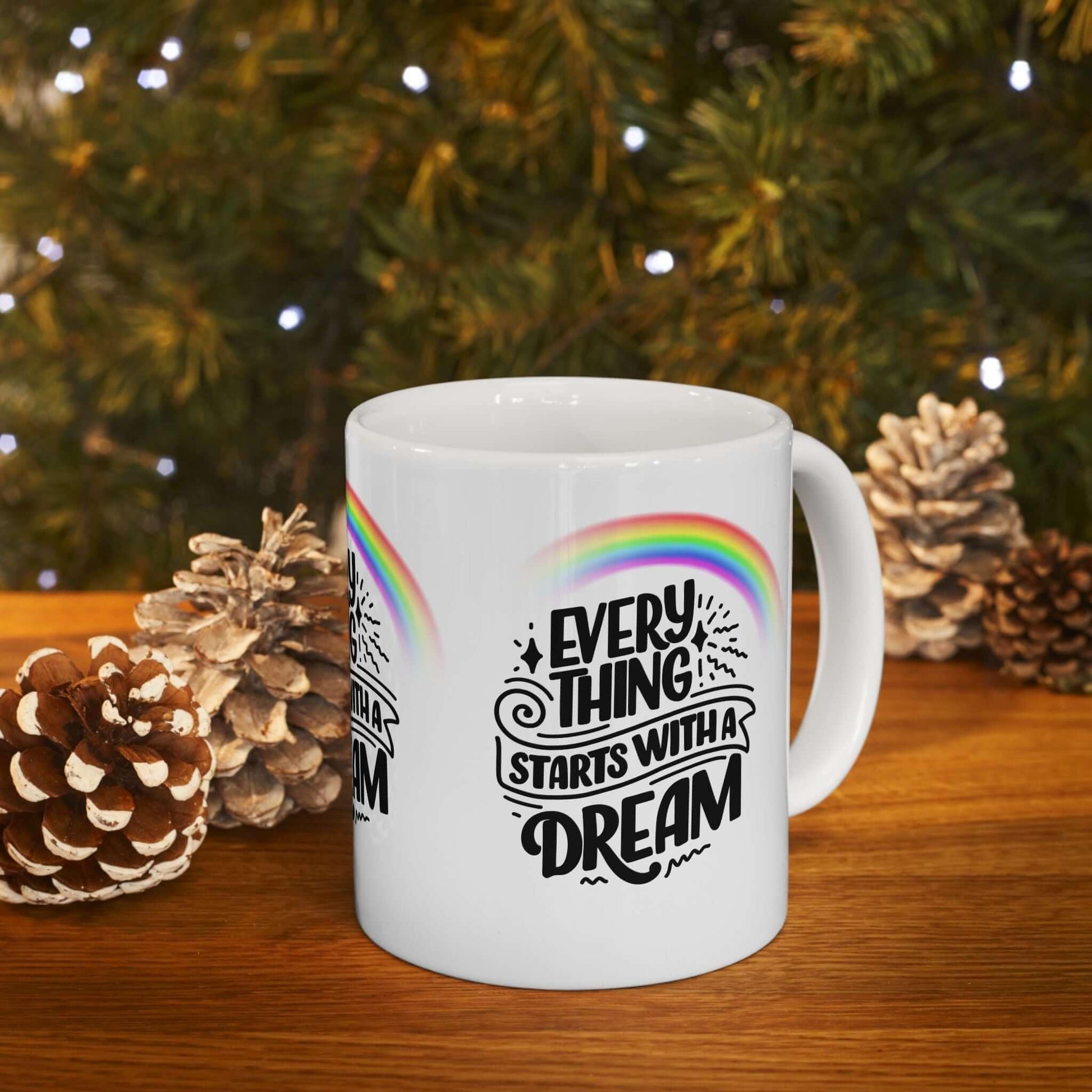 Ceramic coffee mug with rainbow design and 'Everything Starts with a Dream' text, surrounded by pine cones on a wooden surface.