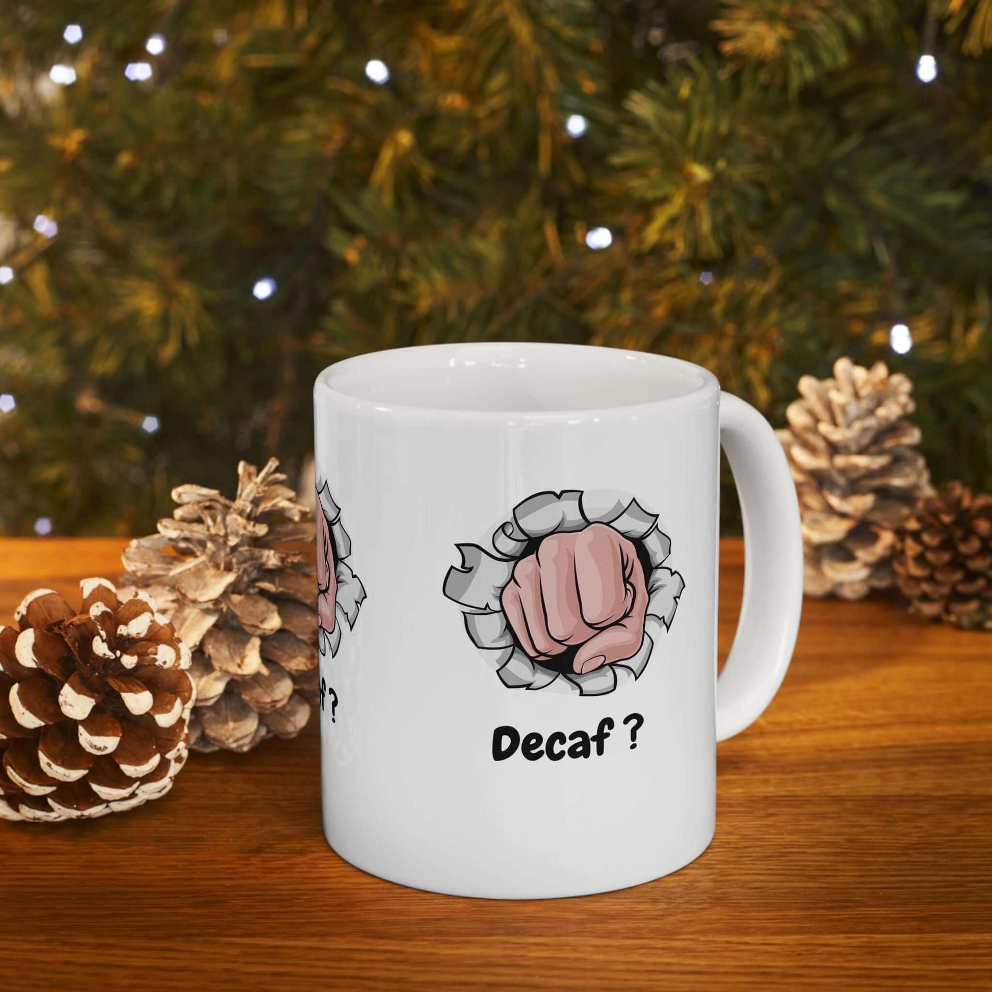 White ceramic mug with 'Decaf?' printed, surrounded by pine cones, on a wooden surface with a green tree background, ideal for coffee lovers.