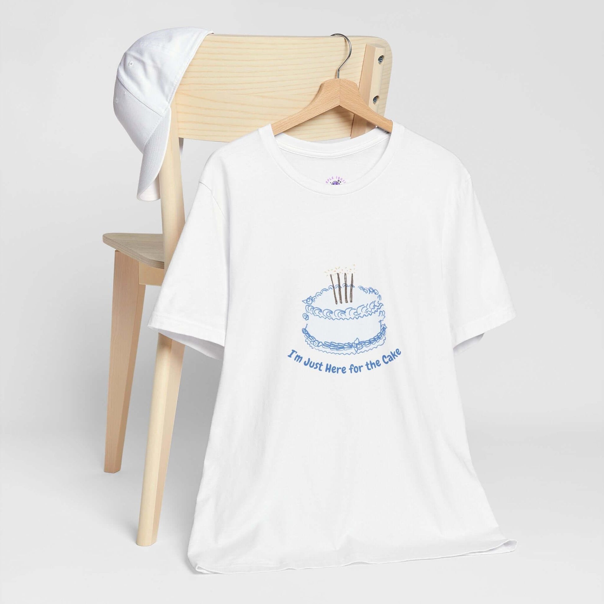 White women's t-shirt with 'I'm Just Here for the Cake' printed on it, featuring a cake graphic, displayed on a wooden chair with a cap.