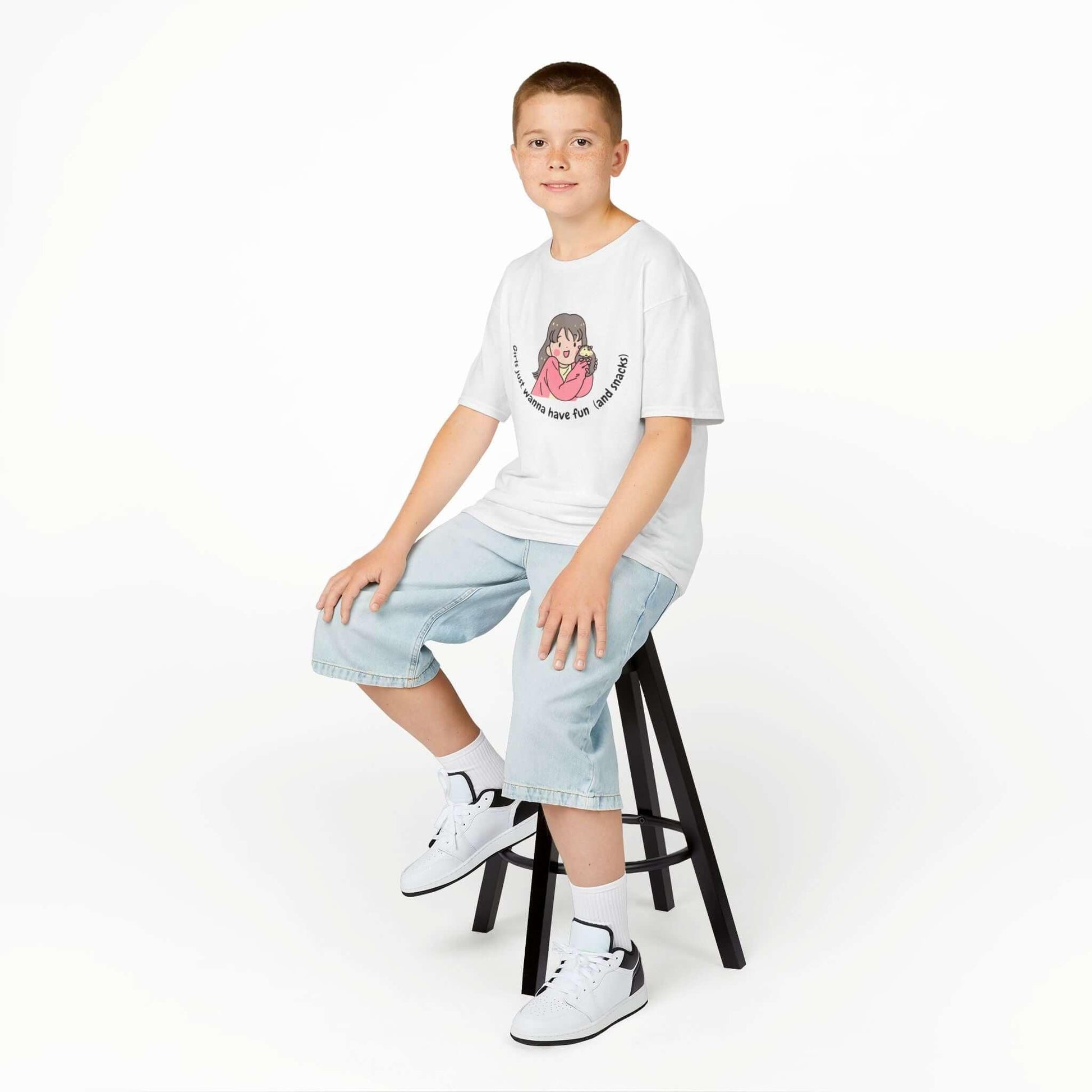 Child wearing a funny kids T-shirt with cartoon graphic, sitting on stool, denim shorts, white sneakers; perfect casual wear.