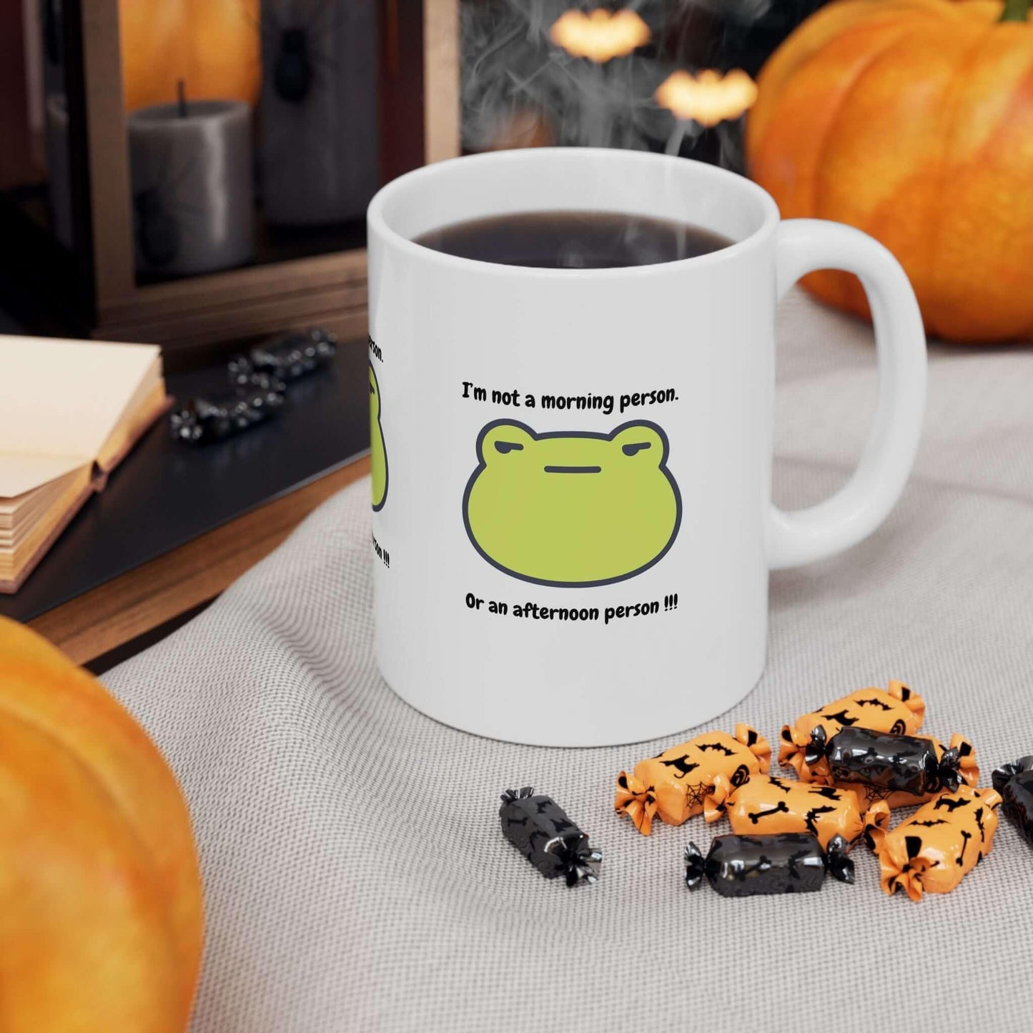 Ceramic coffee mug with funny green frog design and text about morning and afternoon personalities, surrounded by pumpkins and candies.