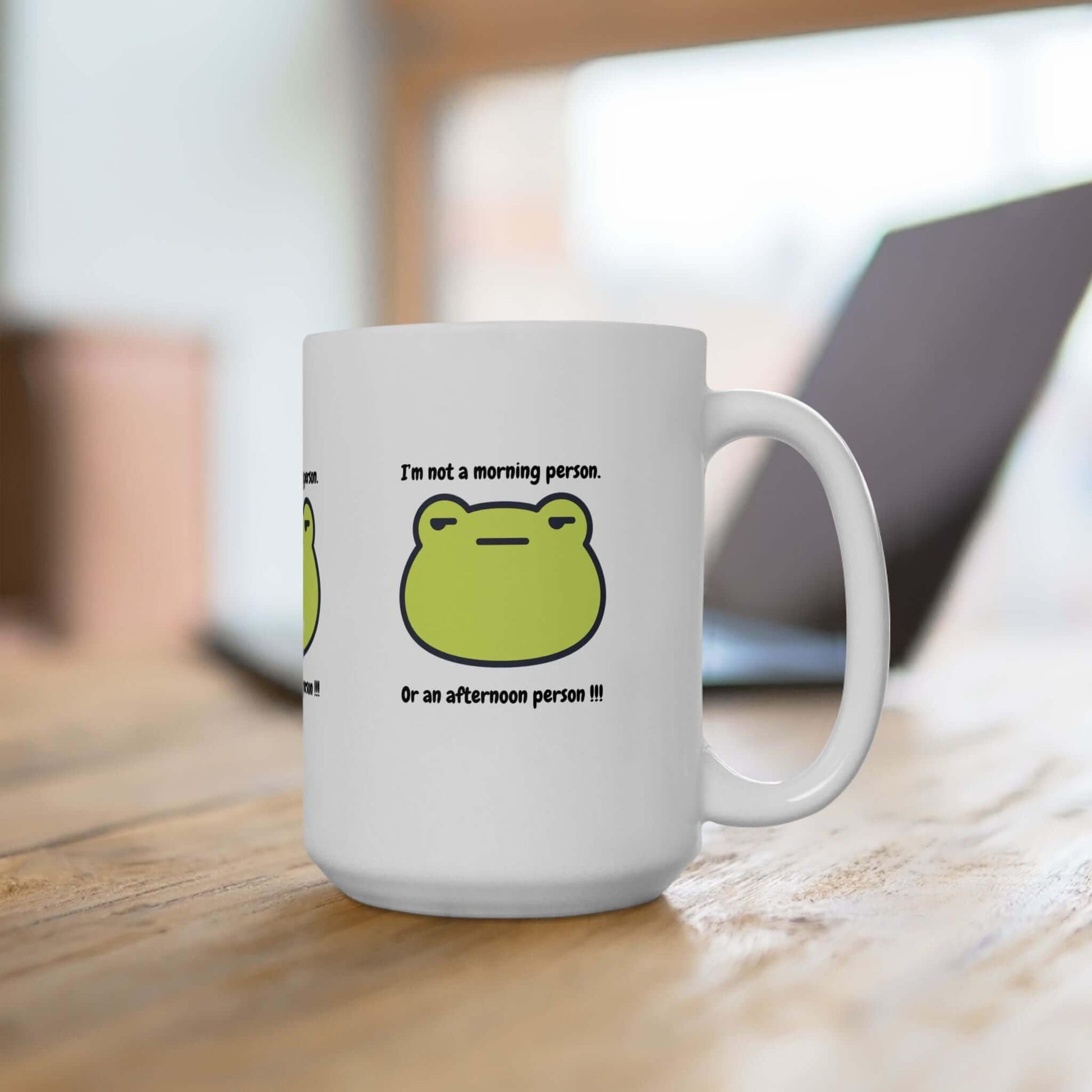 Funny ceramic mug with green frog design and 'I'm not a morning person or an afternoon person' text on a wooden table.