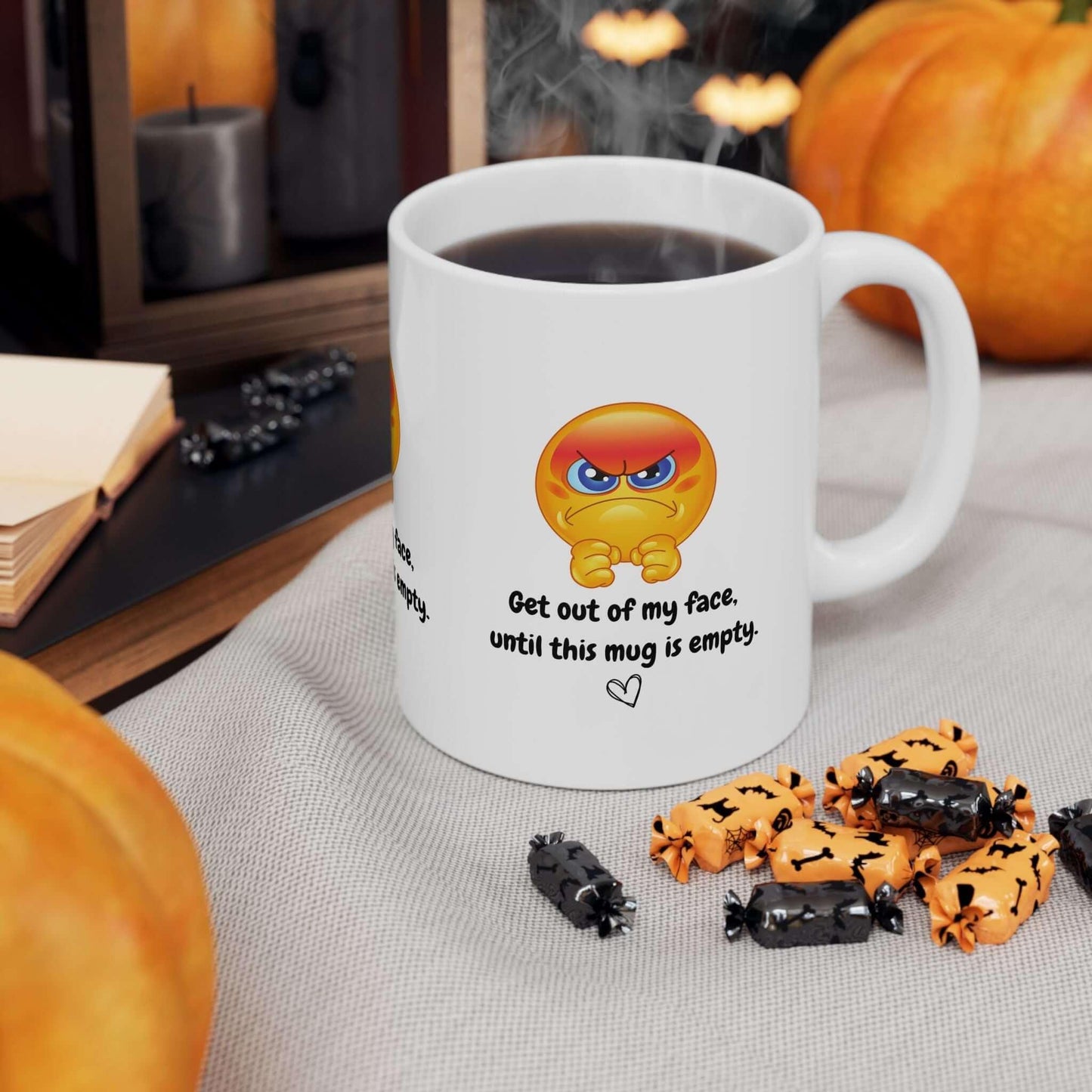 Ceramic mug with angry emoji and 'Get out of my face' text, surrounded by Halloween candy and pumpkins, perfect funny coffee gift.