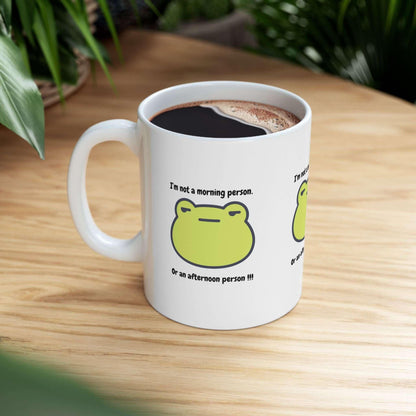 Ceramic mug with "I'm not a morning person. Or an afternoon person" design, placed on a wooden table near green plants.